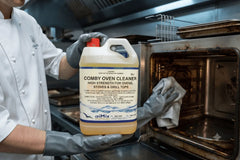 A person in a white chef coat and gray gloves holds a large plastic jug of aiMix Comby Oven Cleaner in front of an open dirty oven in a commercial kitchen setting.