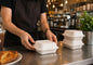 A person in a black apron is shown handling white disposable foam take-out containers on a stainless steel counter in a cafe setting. A small dimension overlay indicates the container is 8 inches wide.