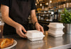 A person in a black apron is shown handling white disposable foam take-out containers on a stainless steel counter in a cafe setting. A small dimension overlay indicates the container is 8 inches wide.