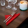 Silverware on a red napkin with glasses and a candle in the background on a wooden table.